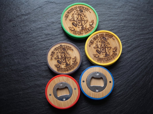 Five round bottle opener coasters arranged on a dark slate surface. Each coaster has a wood-grain center with a metal bottle opener cutout, surrounded by a different rim color: green, grey, yellow, red, and blue.