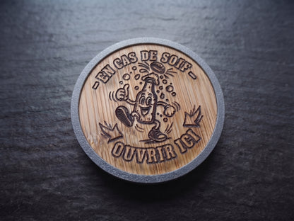 A round wooden opener with a gray textured rim, featuring a laser-engraved cartoon bottle character and the French text 'EN CAS DE SOIF - OUVRIR ICI' on a dark slate background.