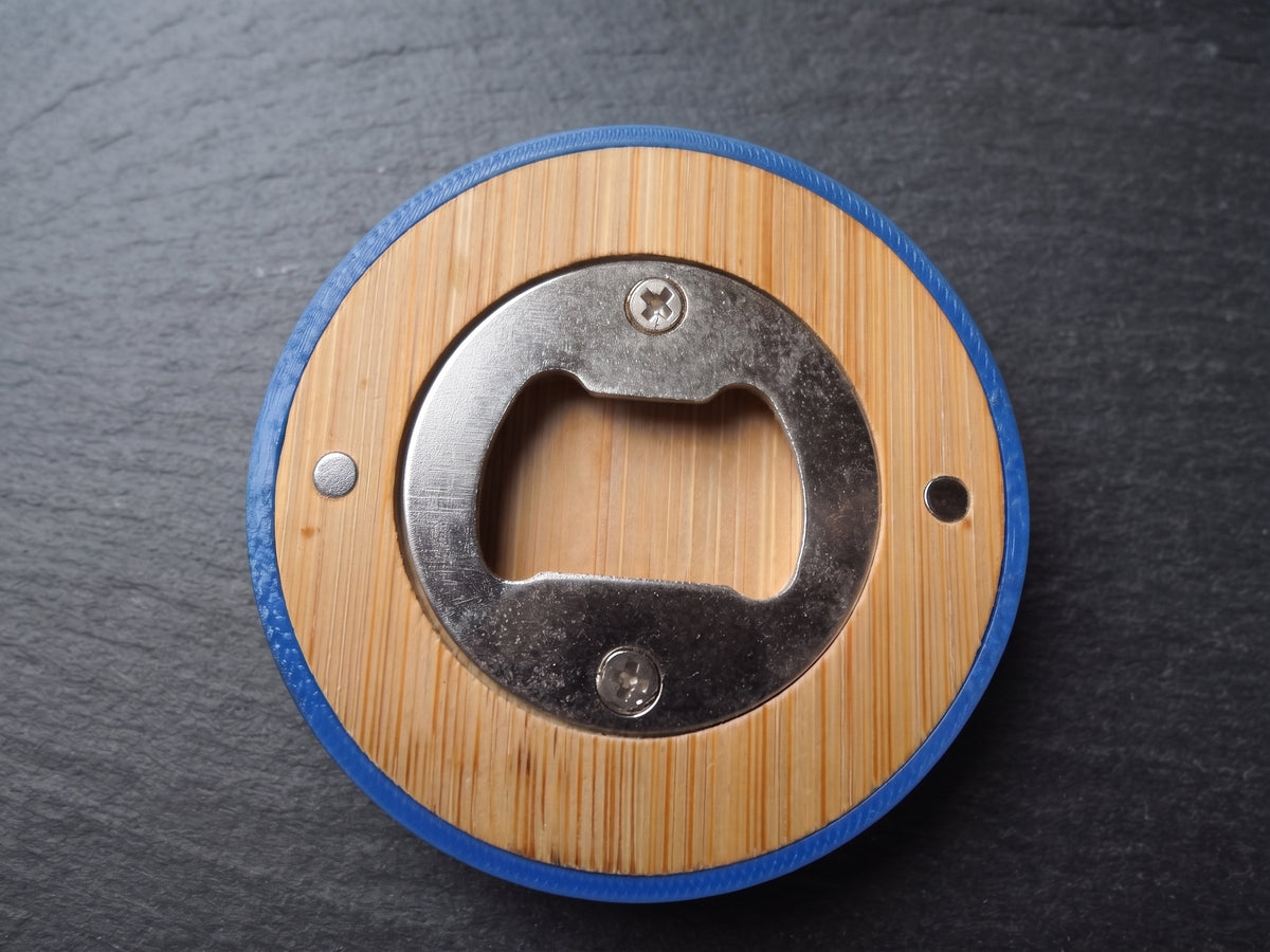 A circular bottle opener with a light wood grain center, a blue outer ring, and a silver metal opener mechanism, shown on a dark slate background.