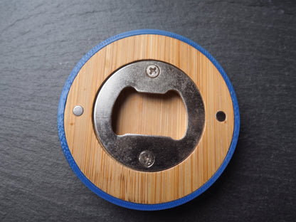 A circular bottle opener with a light wood grain center, a blue outer ring, and a silver metal opener mechanism, shown on a dark slate background.