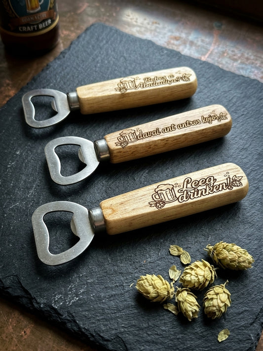 Three wooden-handled bottle openers with metal pull tabs are arranged on a dark slate board, each engraved with a beer-themed phrase. A bottle cap and dried hops are nearby, with a partial beer bottle visible in the upper left corner.