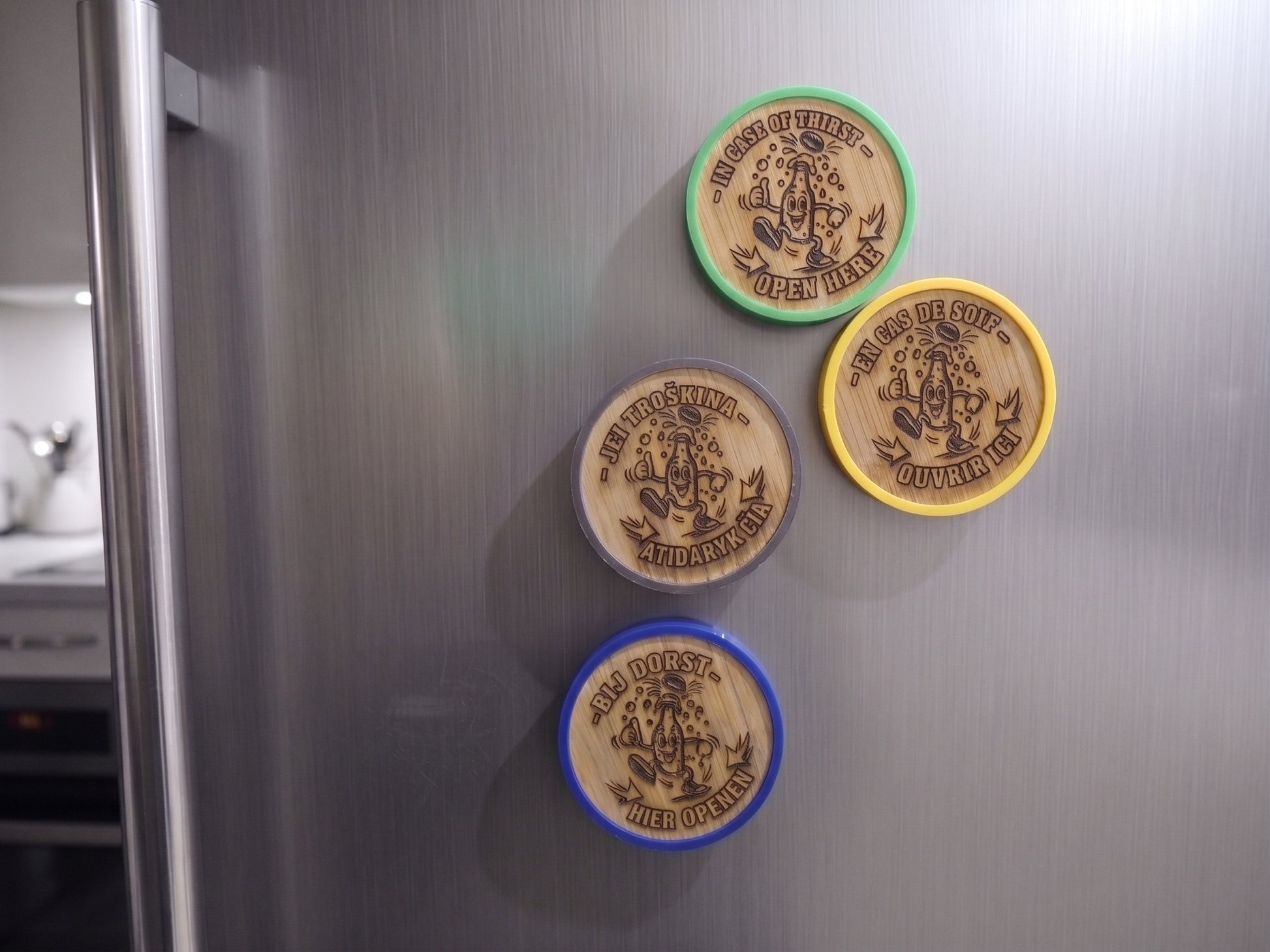 Four round wooden magnets with engraved designs and colorful borders (green, grey, yellow, and blue) attached to a brushed stainless steel refrigerator door.
