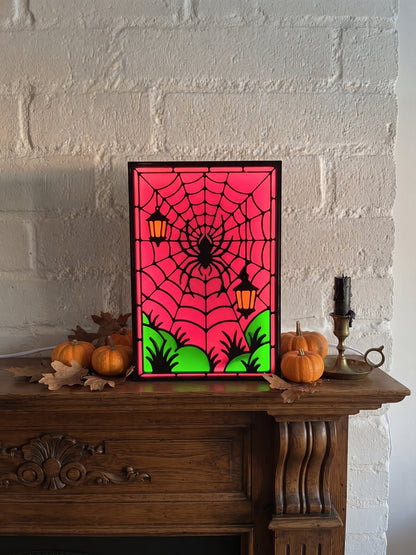 A framed Halloween stained-glass-style art piece with a magenta spiderweb background, black spider and cobwebs, green grass, and orange lanterns, displayed on a wooden mantel with pumpkins and a candle against a white brick wall.