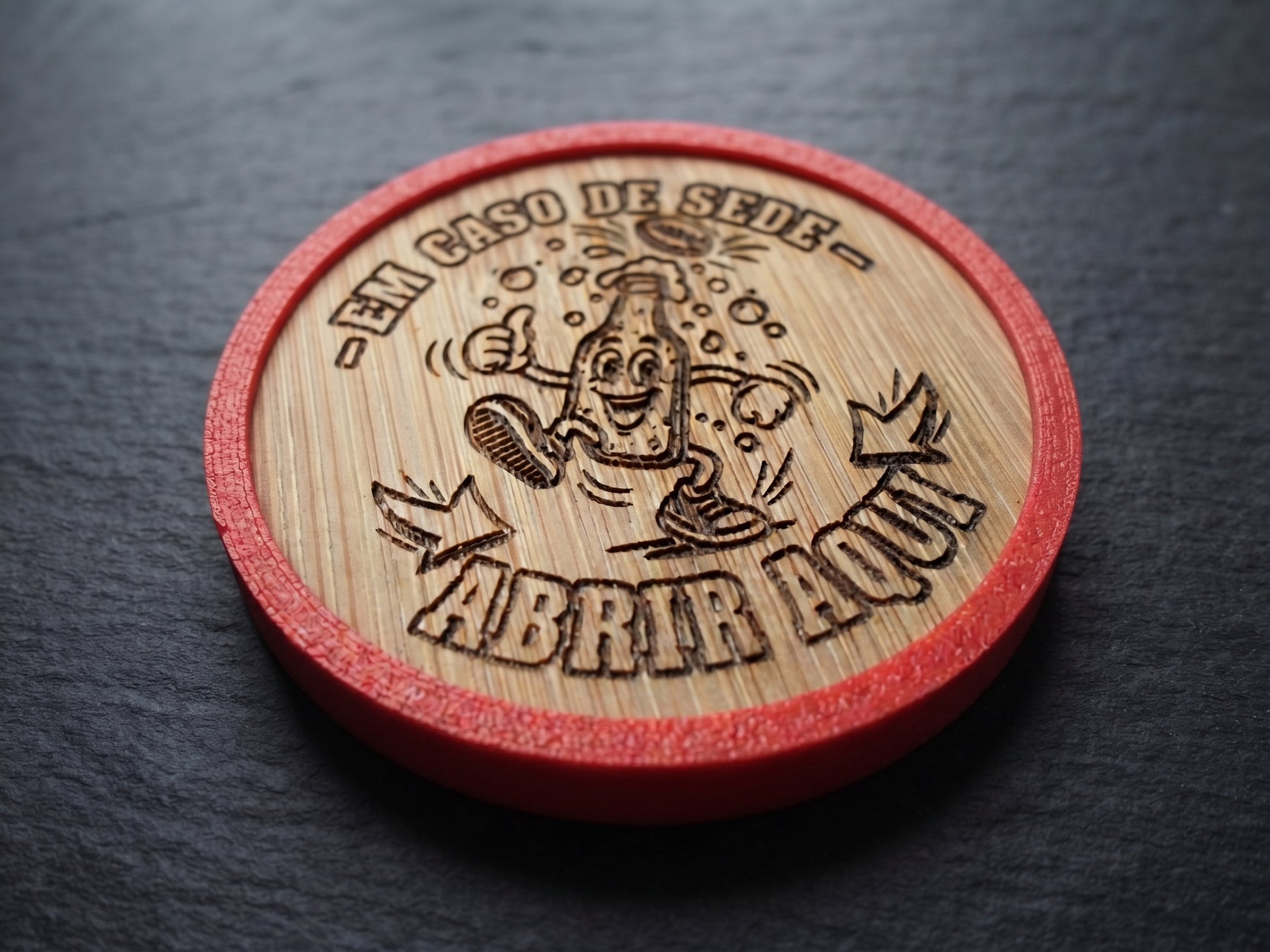 A round wooden bottle-cap-style opener with a red outer rim, photographed on a dark slate surface. The natural wood top is engraved with a cartoon bottle character and Portuguese text reading “-EM CASO DE SEDE- ABRIR AQUI.