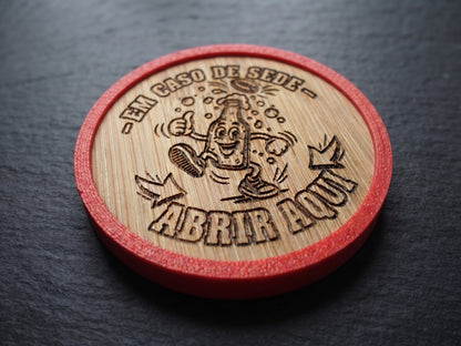 A round wooden bottle-cap-style opener with a red outer rim, photographed on a dark slate surface. The natural wood top is engraved with a cartoon bottle character and Portuguese text reading “-EM CASO DE SEDE- ABRIR AQUI.