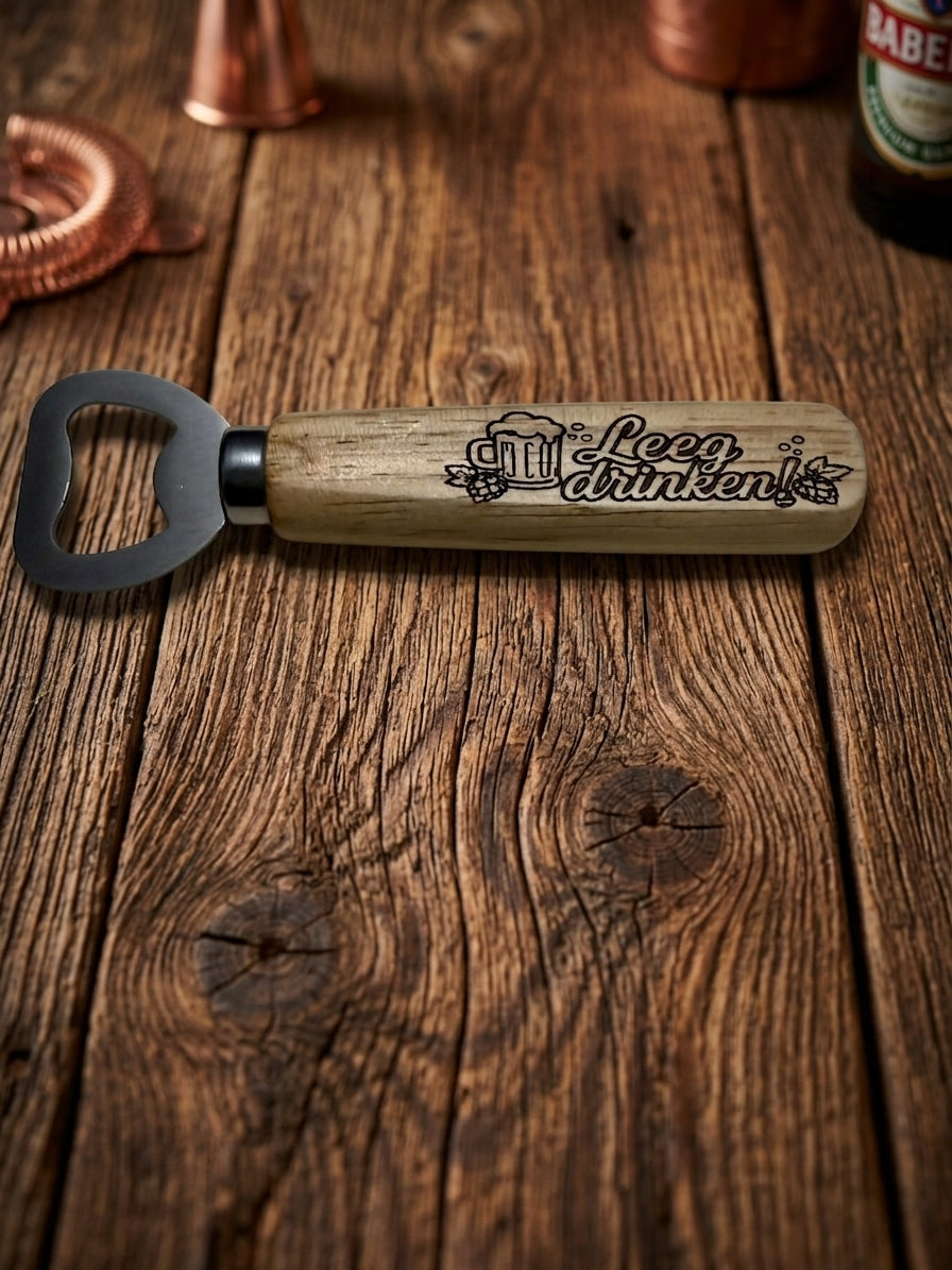A wooden-handled bottle opener with a metal blade, engraved with 'Leeg drinken' and beer-themed illustrations, resting on a rustic wooden tabletop with copper bar tools and a bottle partially visible in the background.