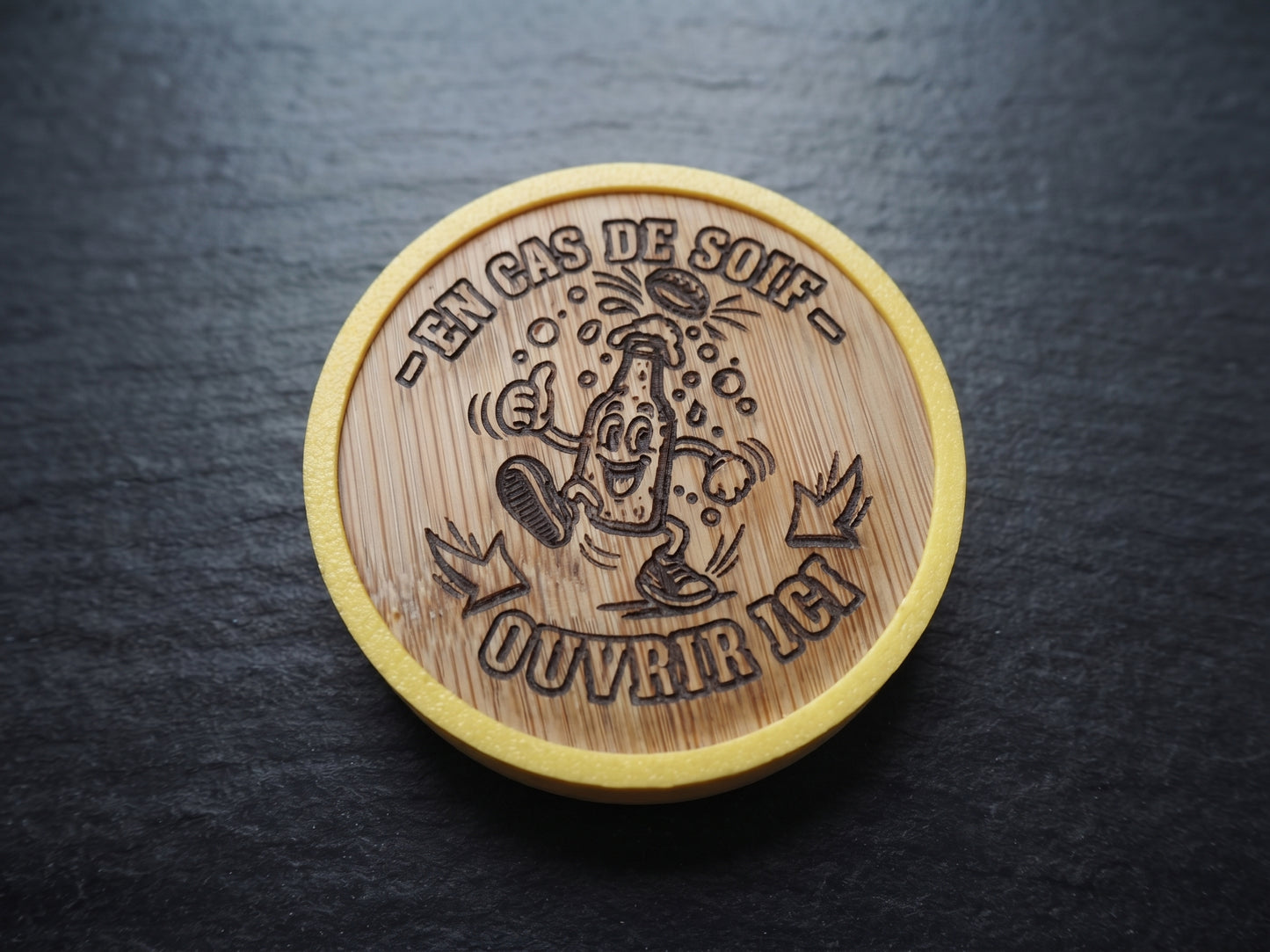 A round wooden opener with a natural bamboo texture and a yellow painted rim, featuring a cartoon illustration of a drinking glass and the French text '-EN CAS DE SOIF - OUVRIR ICI' engraved in the center, placed on a dark slate surface.