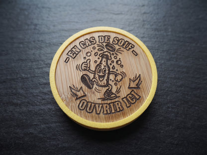 A round wooden opener with a natural bamboo texture and a yellow painted rim, featuring a cartoon illustration of a drinking glass and the French text '-EN CAS DE SOIF - OUVRIR ICI' engraved in the center, placed on a dark slate surface.
