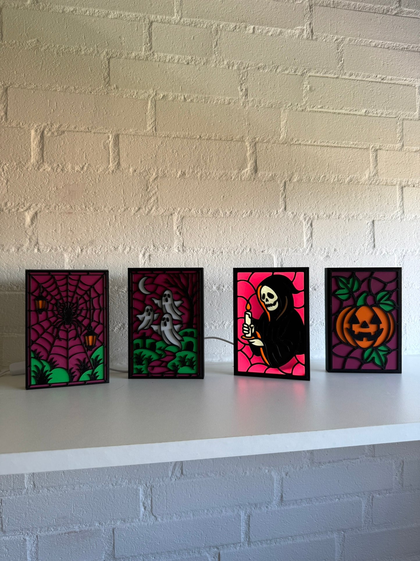 Four Halloween-themed art panels displayed on a white shelf against a white brick wall. The panels feature a spiderweb, a grim reaper, a ghost under a crescent moon, and a pumpkin.