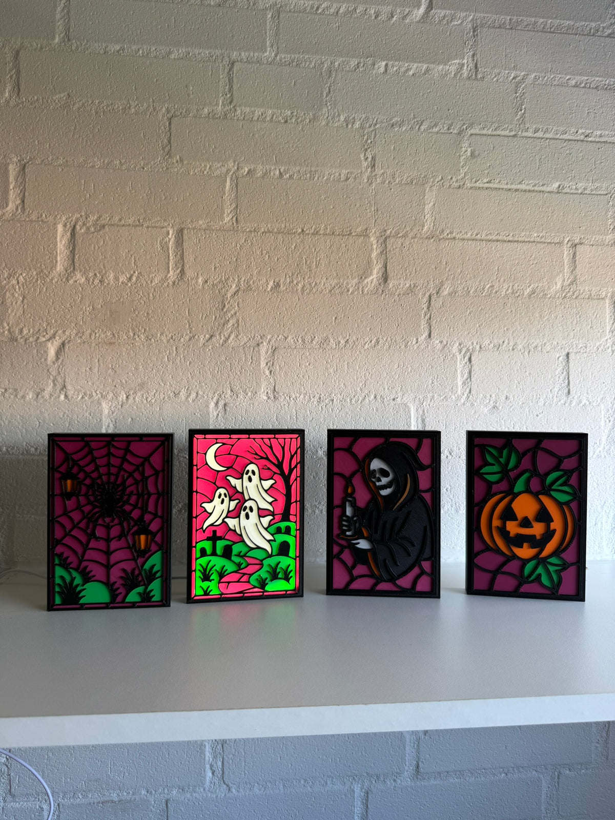 Four small framed Halloween art prints displayed side-by-side on a white shelf against a white brick wall. The prints feature classic spooky designs: a spiderweb, ghosts under a crescent moon, the Grim Reaper, and a carved pumpkin, all in a white, magenta, green, orange and black stained-glass style.