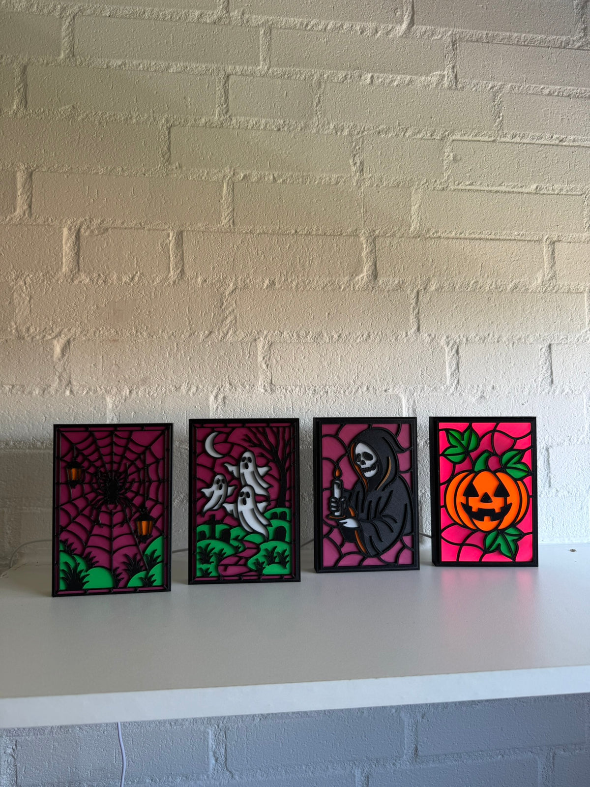 Four small framed Halloween art prints displayed side-by-side on a white shelf against a white painted brick wall. The prints feature a spiderweb, ghosts, the grim reaper, and a pumpkin in a stained-glass style with magenta backgrounds.