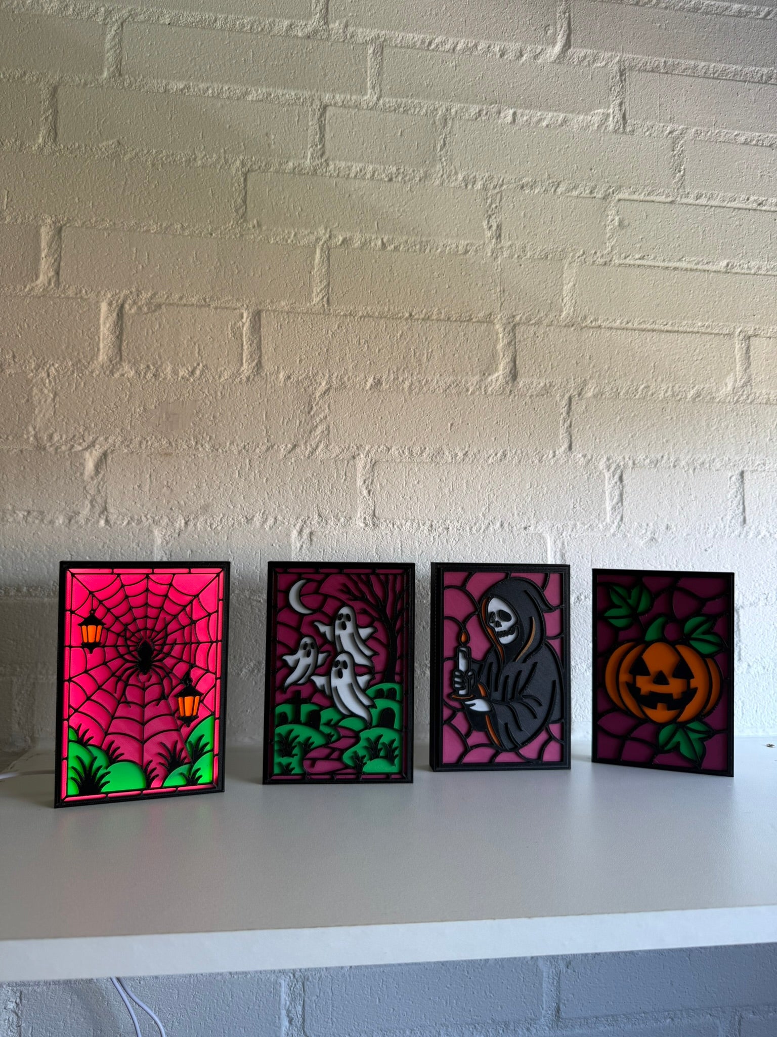 Four small framed Halloween art prints displayed side-by-side on a white shelf against a white brick wall. The prints feature vibrant stained-glass-style designs: a spiderweb with pumpkins, ghosts in a haunted house, a skeleton, and a pumpkin.