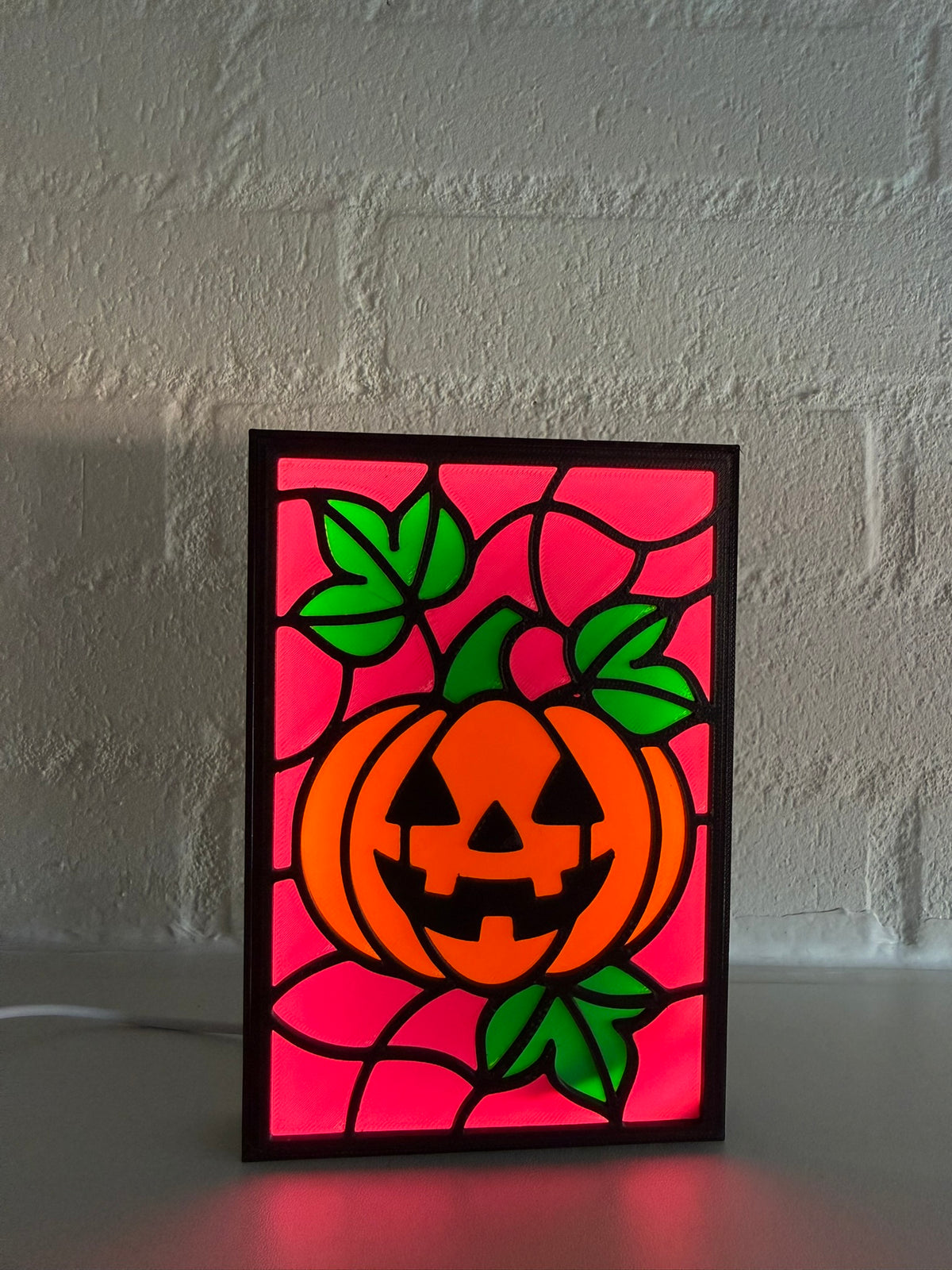 A rectangular stained-glass-style Halloween sign featuring a glowing orange pumpkin with green leaves against a magenta background, standing on a surface in front of a light brick wall.
