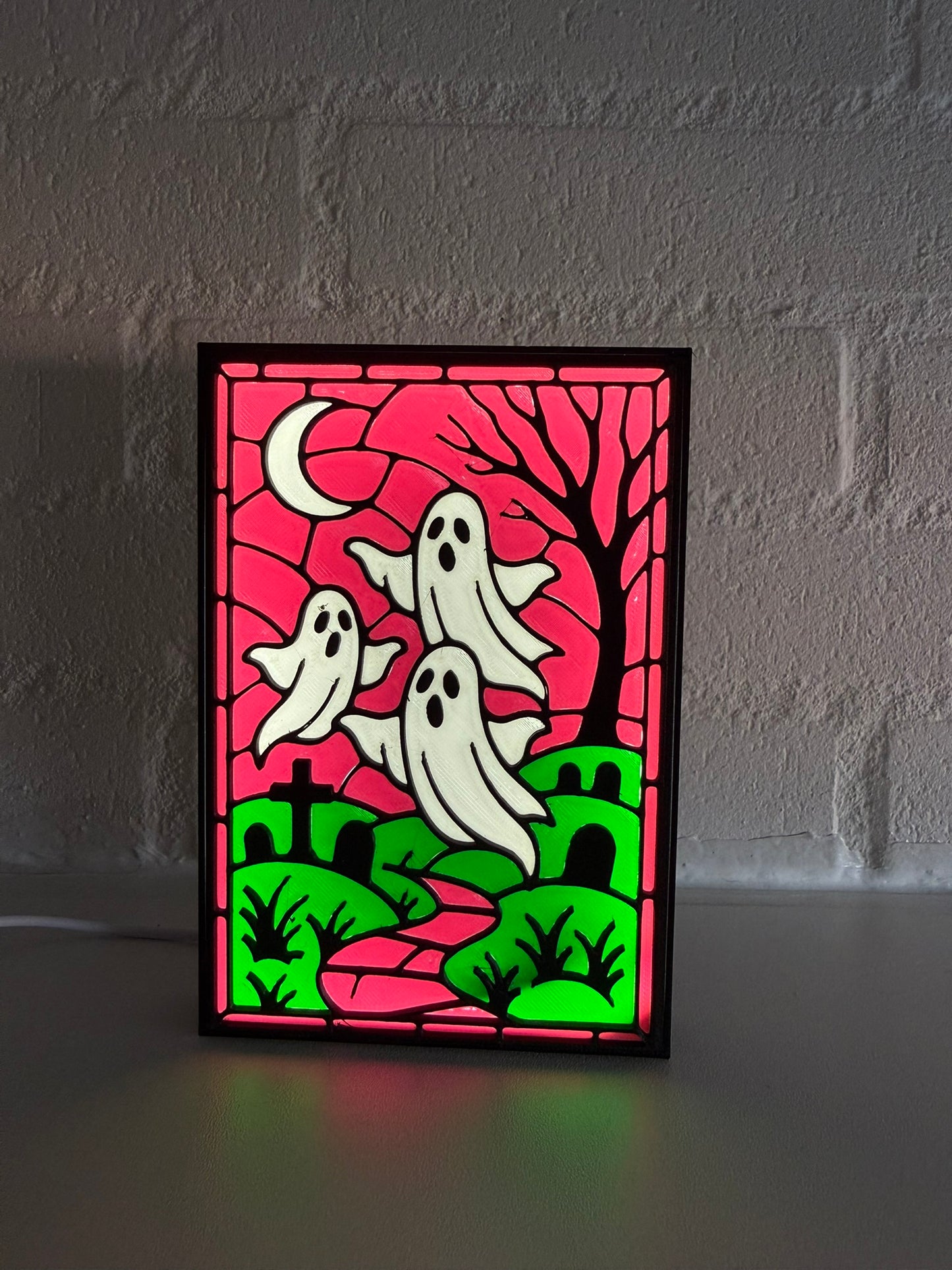 A rectangular Halloween stained-glass style light or panel with three white ghosts, a black tree, green hills, and a magenta sky, standing on a surface against a gray brick wall.