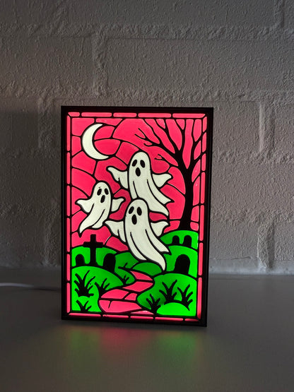 A rectangular Halloween stained-glass style light or panel with three white ghosts, a black tree, green hills, and a magenta sky, standing on a surface against a gray brick wall.
