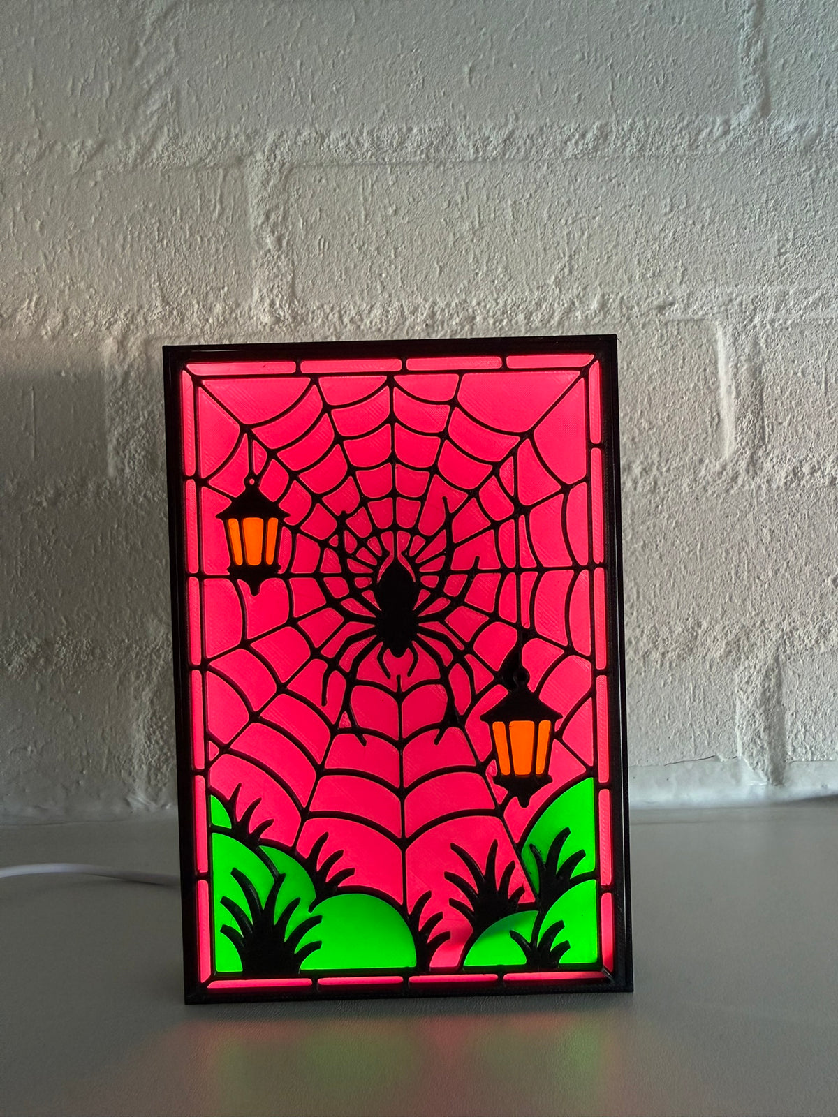 A framed stained-glass-style Halloween sign featuring a black spider in a web, magenta background, green grass, and two orange lanterns, displayed against a white brick wall.