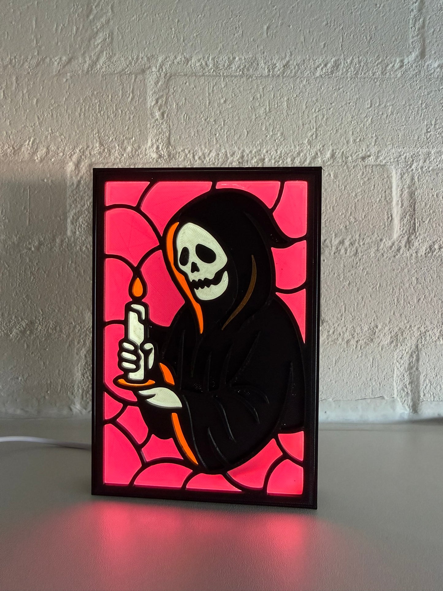 A framed art print featuring a grim reaper with a skull face and black hood holding a lit candle, styled like stained glass with a magenta background, displayed on a white surface against a white brick wall.