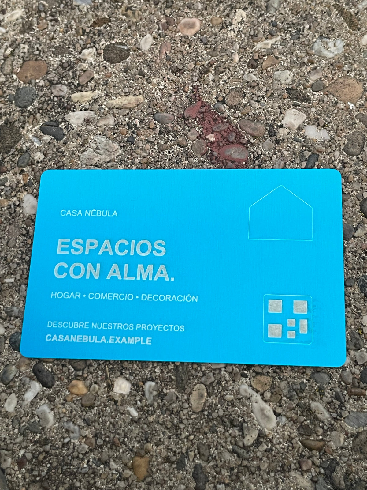 A bright blue card with white Spanish text reading 'CASA NÉBULA' and 'ESPACIOS CON ALMA.' placed on a textured gravel surface.