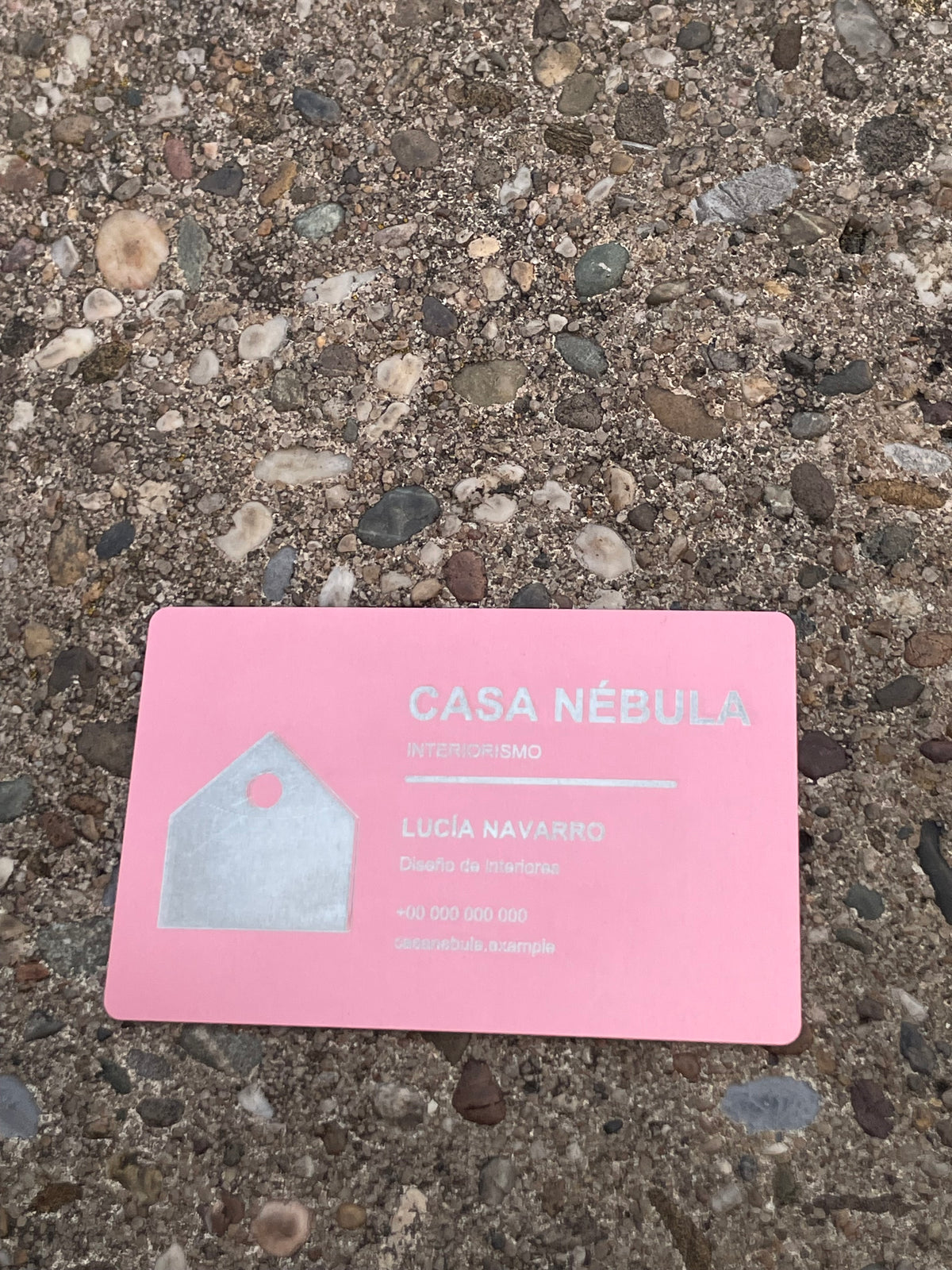 A light pink business card with text reading 'CASA NÉBULA' and 'Lucía Navarro' lies on a textured concrete surface made of small pebbles and gravel.