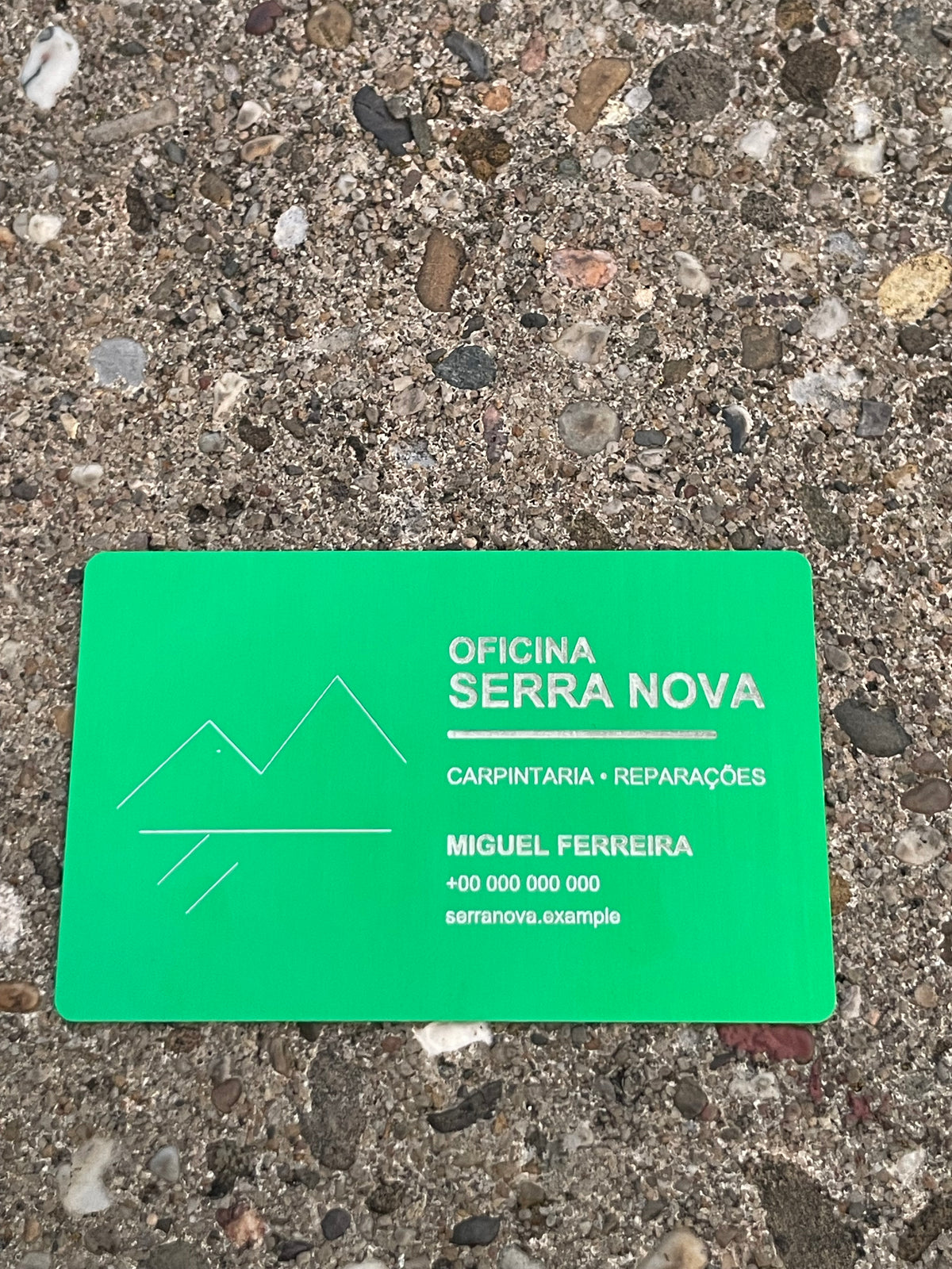 A bright green rectangular business card with engraved text and a simple mountain-and-water logo, placed on a rough concrete surface. The card reads 'Oficina Serra Nova', 'Carpintaria - Reparações', and 'Miguel Ferreira' with a phone number.