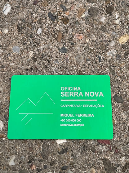 A bright green rectangular business card with engraved text and a simple mountain-and-water logo, placed on a rough concrete surface. The card reads 'Oficina Serra Nova', 'Carpintaria - Reparações', and 'Miguel Ferreira' with a phone number.