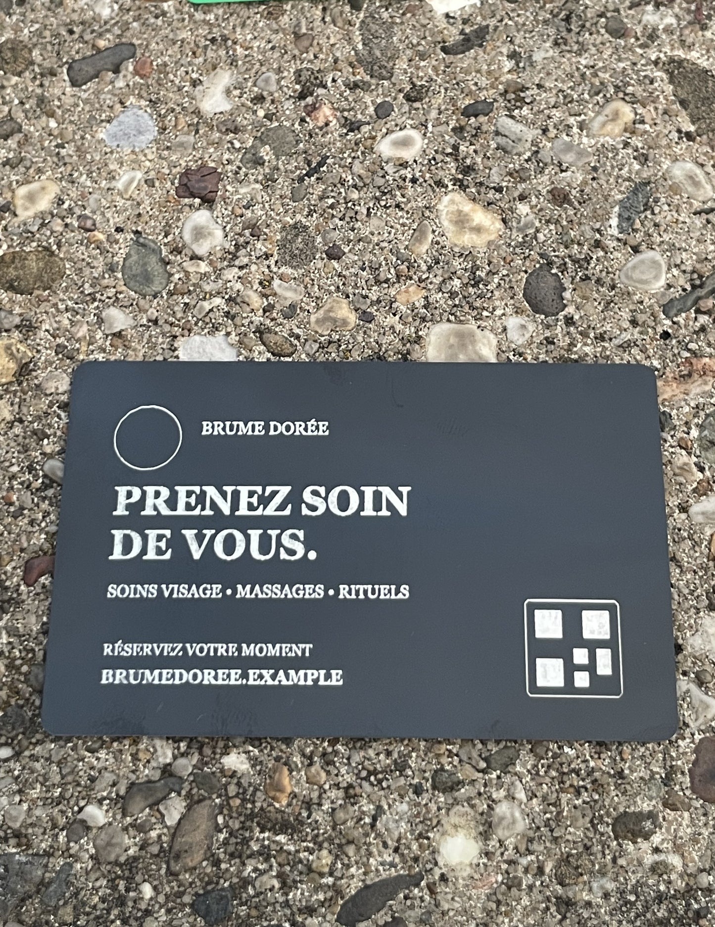 A dark business card with French text reading 'PRENEZ SOIN DE VOUS.' and 'BRUME DORÉE,' placed on a gravel surface outdoors.
