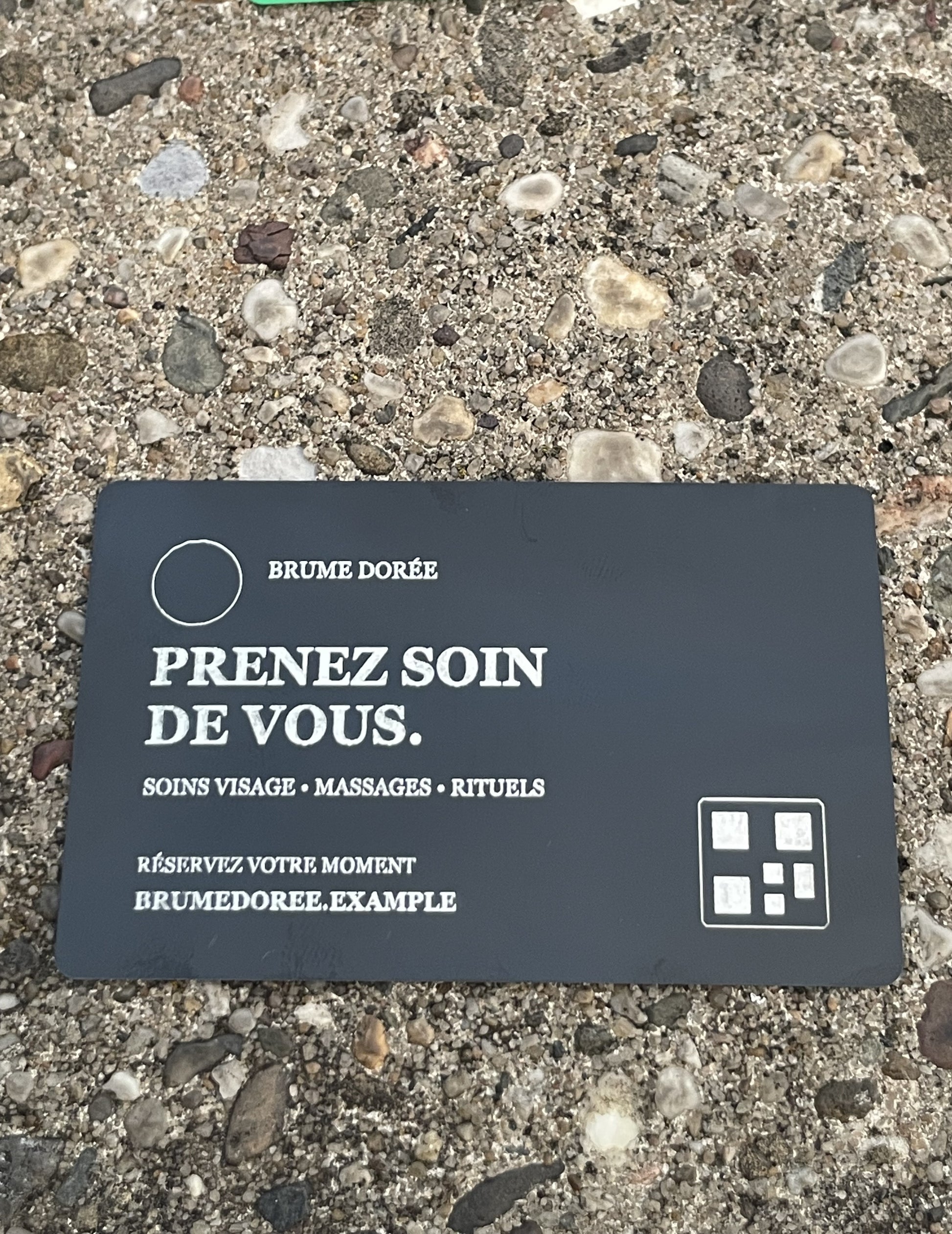 A dark business card with French text reading 'PRENEZ SOIN DE VOUS.' and 'BRUME DORÉE,' placed on a gravel surface outdoors.