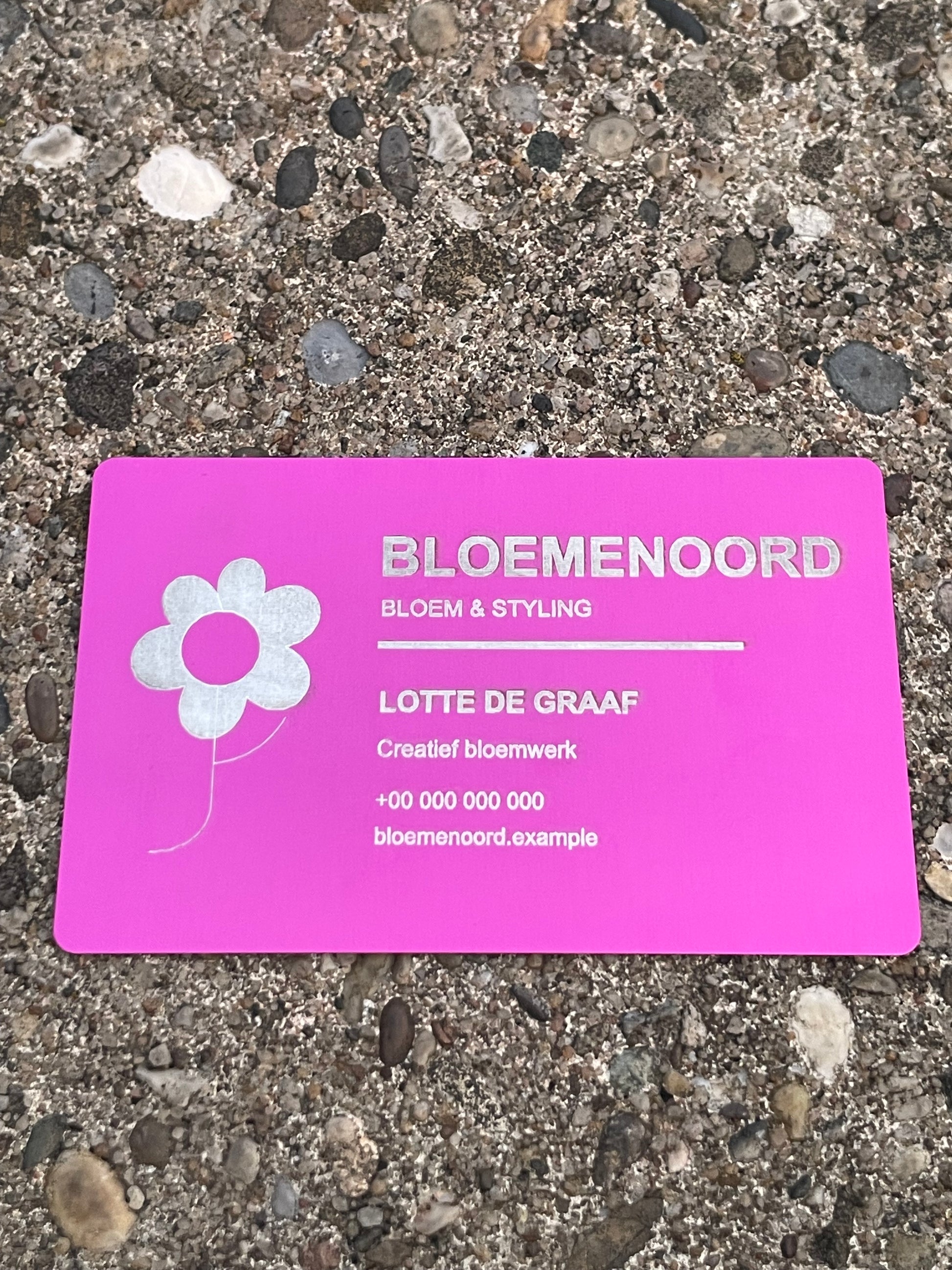 A pink business card for Bloemenoord Bloem & Styling lies flat on a textured gray pebble surface. The card features a white flower graphic on the left and silver foil text with the name Lotte de Graaf.