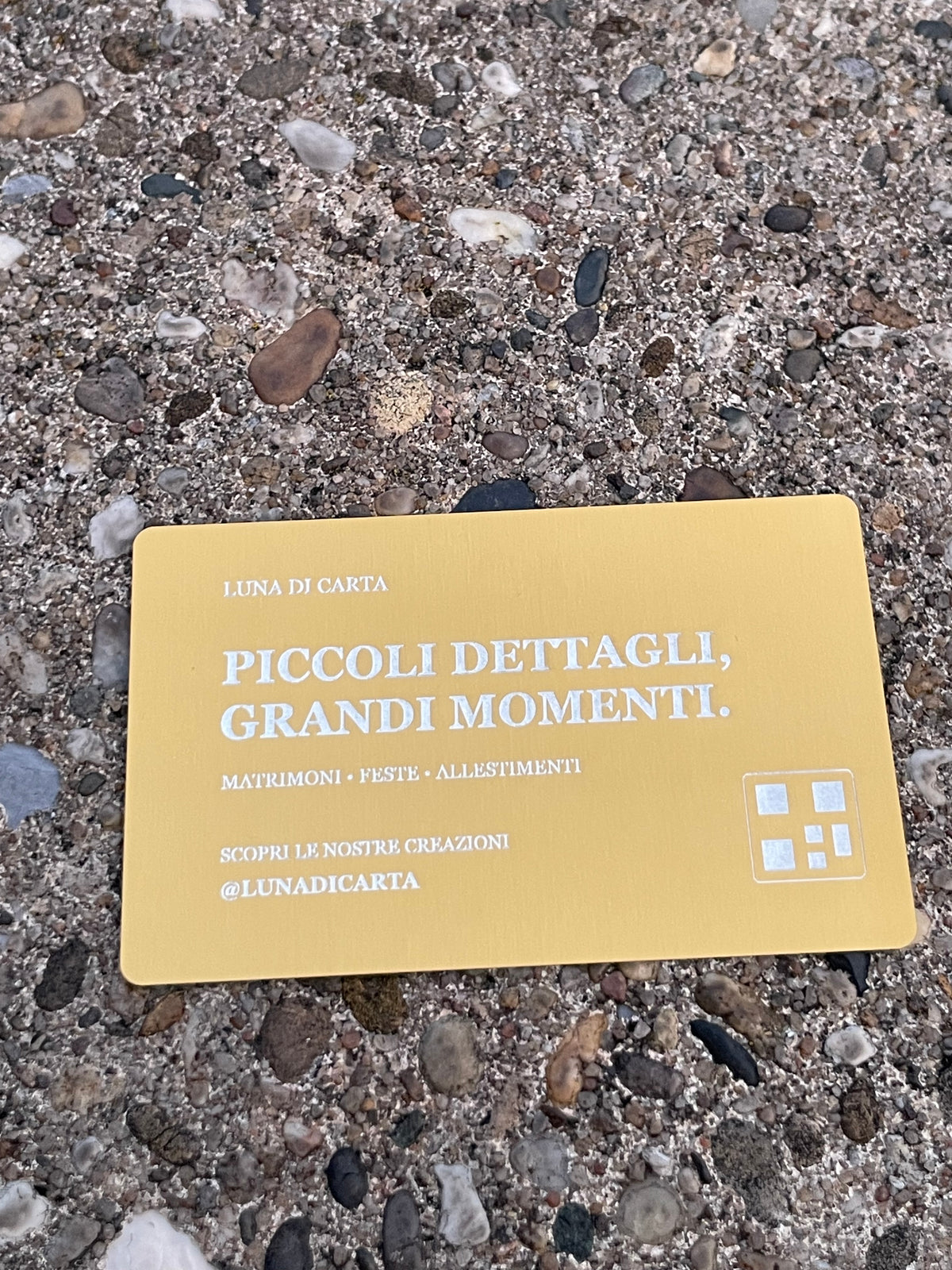 A rectangular gold business card with Italian text lies on a textured concrete surface with small stones and pebbles.