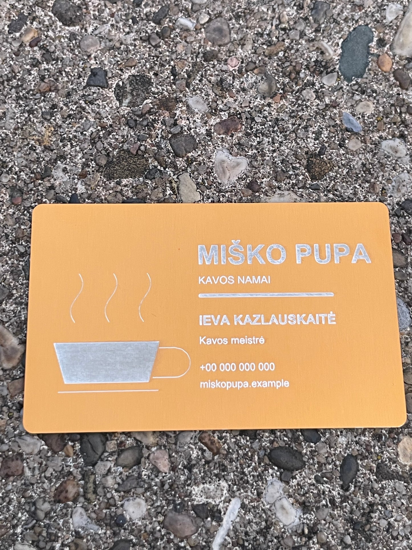A rectangular orange card with Lithuanian text and a simple coffee cup illustration, placed on a gravel surface. The card reads “MIŠKO PUPA” and appears to be the product being shown.