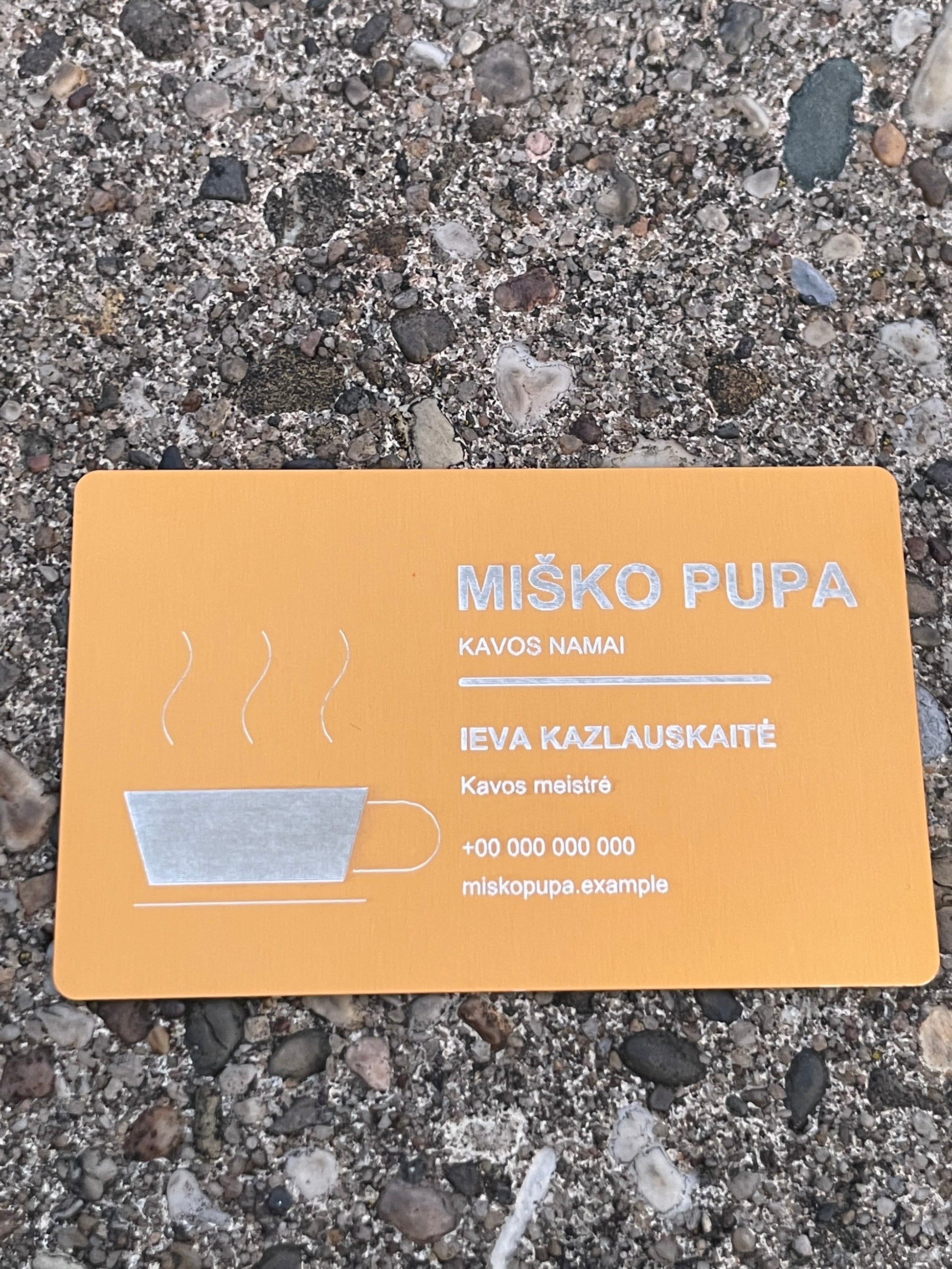 A rectangular orange card with Lithuanian text and a simple coffee cup illustration, placed on a gravel surface. The card reads “MIŠKO PUPA” and appears to be the product being shown.
