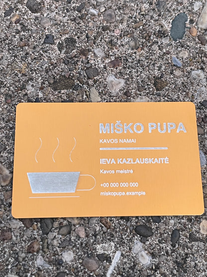 A rectangular orange card with Lithuanian text and a simple coffee cup illustration, placed on a gravel surface. The card reads “MIŠKO PUPA” and appears to be the product being shown.
