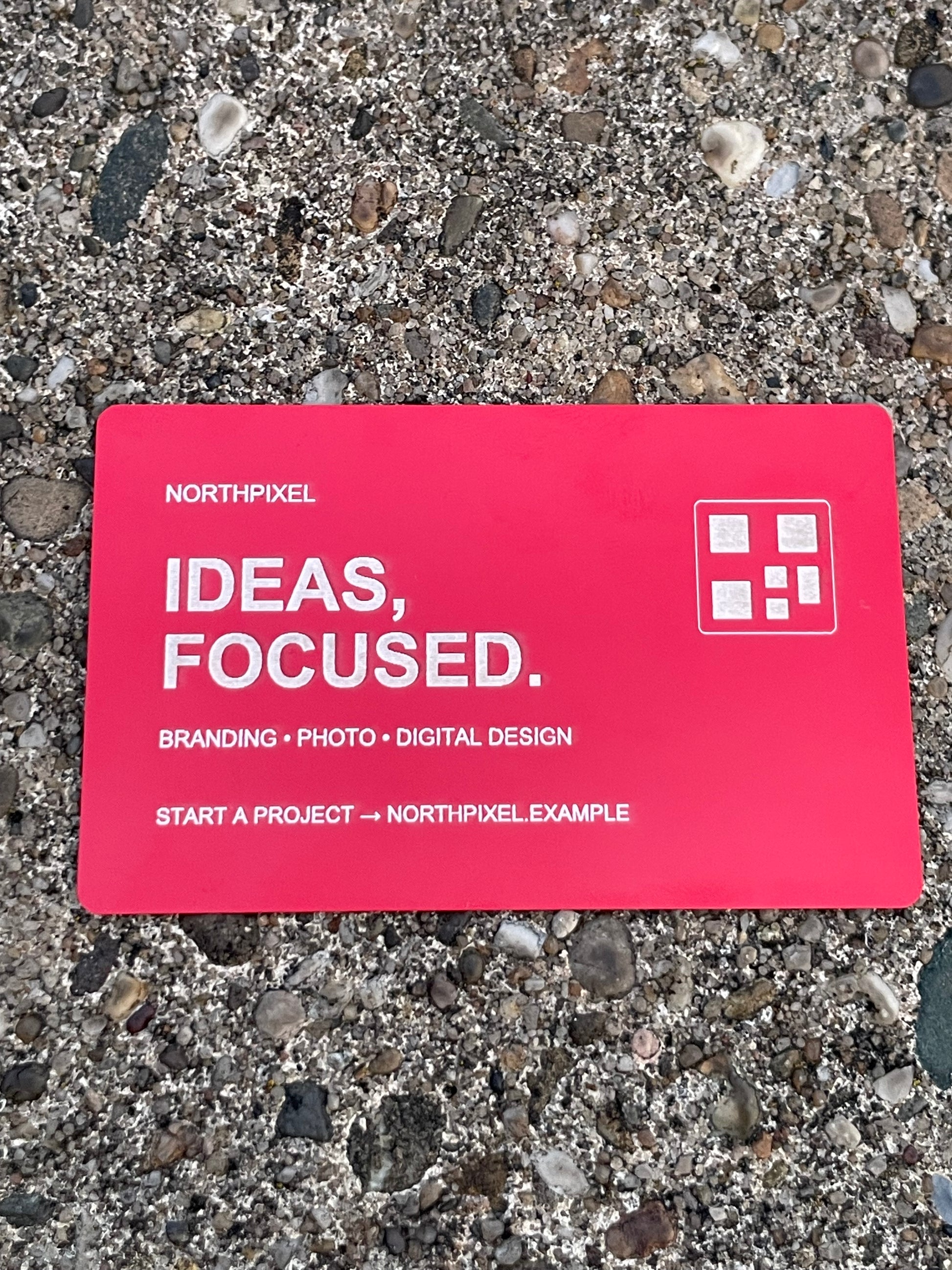 A red business card with text reading 'NORTHPIXEL IDEAS, FOCUSED.' and a small white logo, placed on a gravel surface.