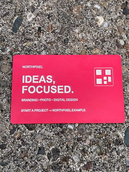 A red business card with text reading 'NORTHPIXEL IDEAS, FOCUSED.' and a small white logo, placed on a gravel surface.