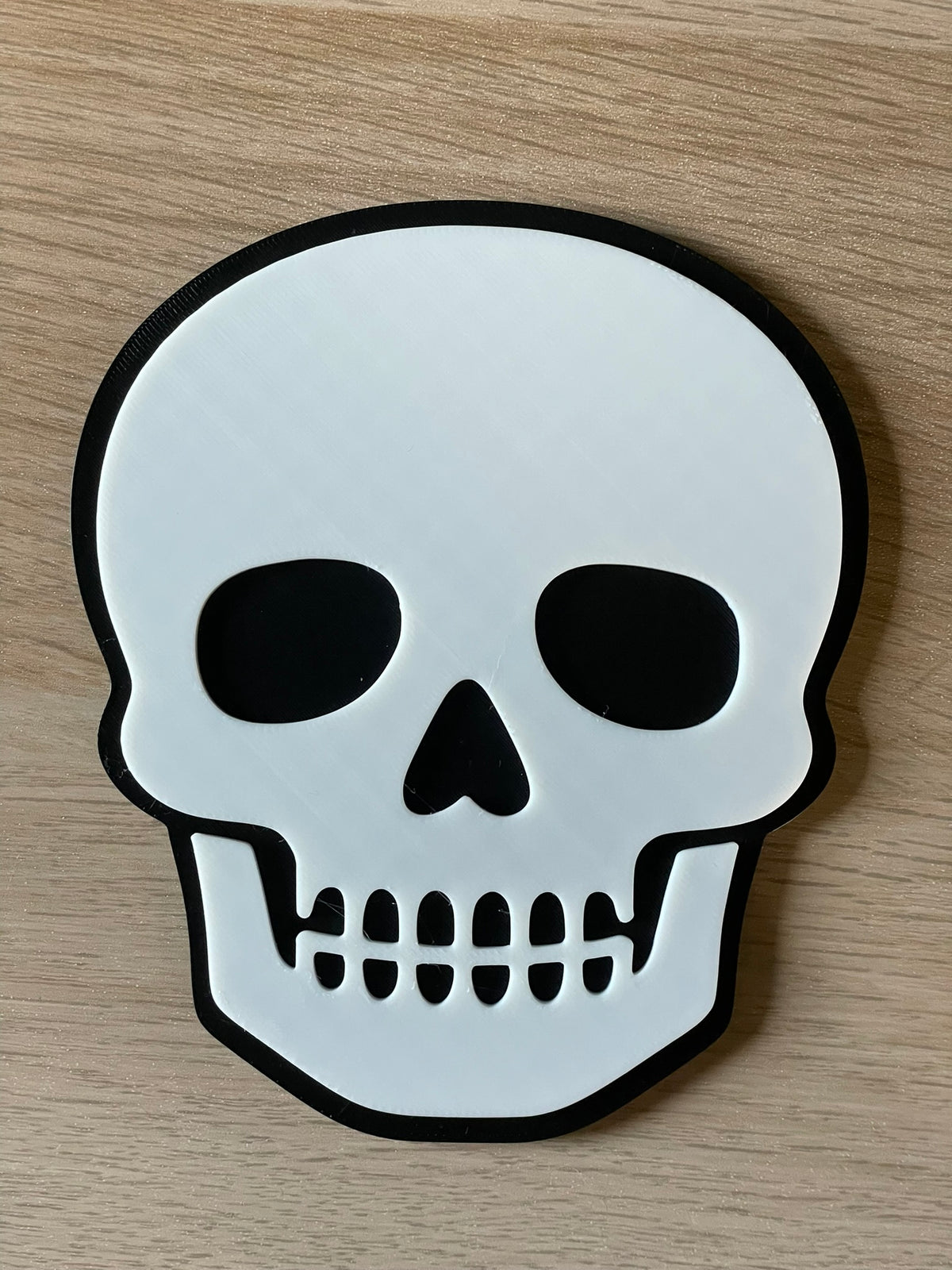 A white skull-shaped coaster with a black outline, photographed flat on a light wood surface.