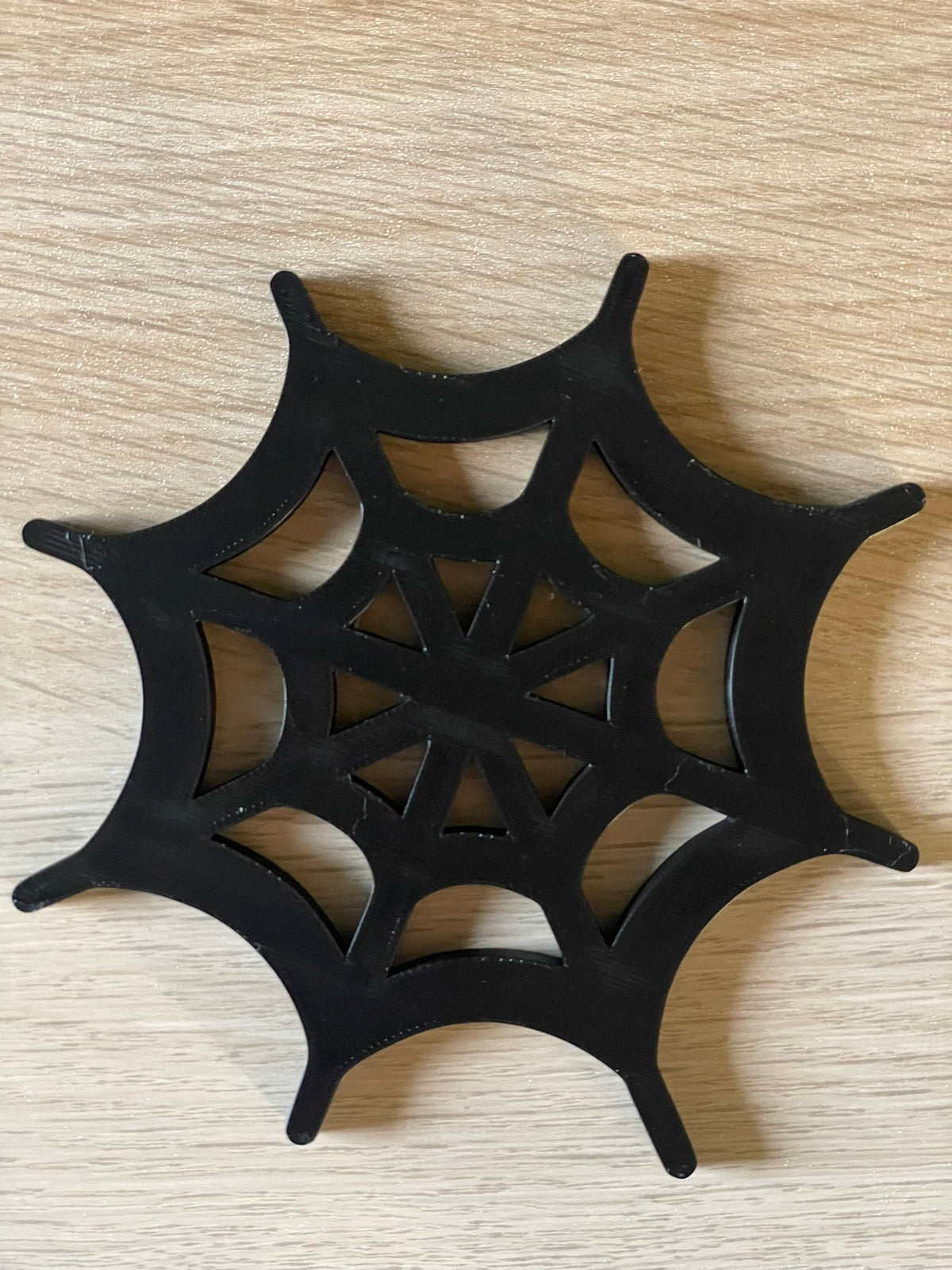 A black spiderweb-shaped decorative coaster with a cutout web pattern, placed on a light wood-grain surface.