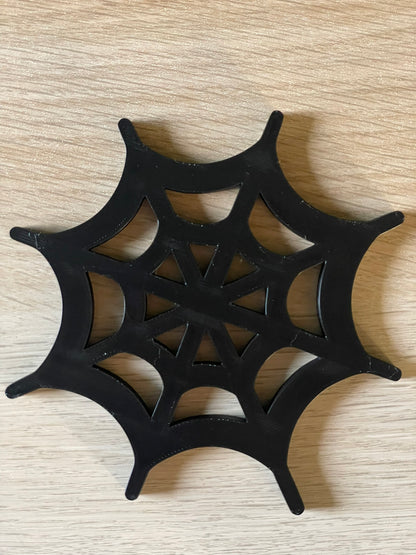 A black spiderweb-shaped decorative coaster with a cutout web pattern, placed on a light wood-grain surface.