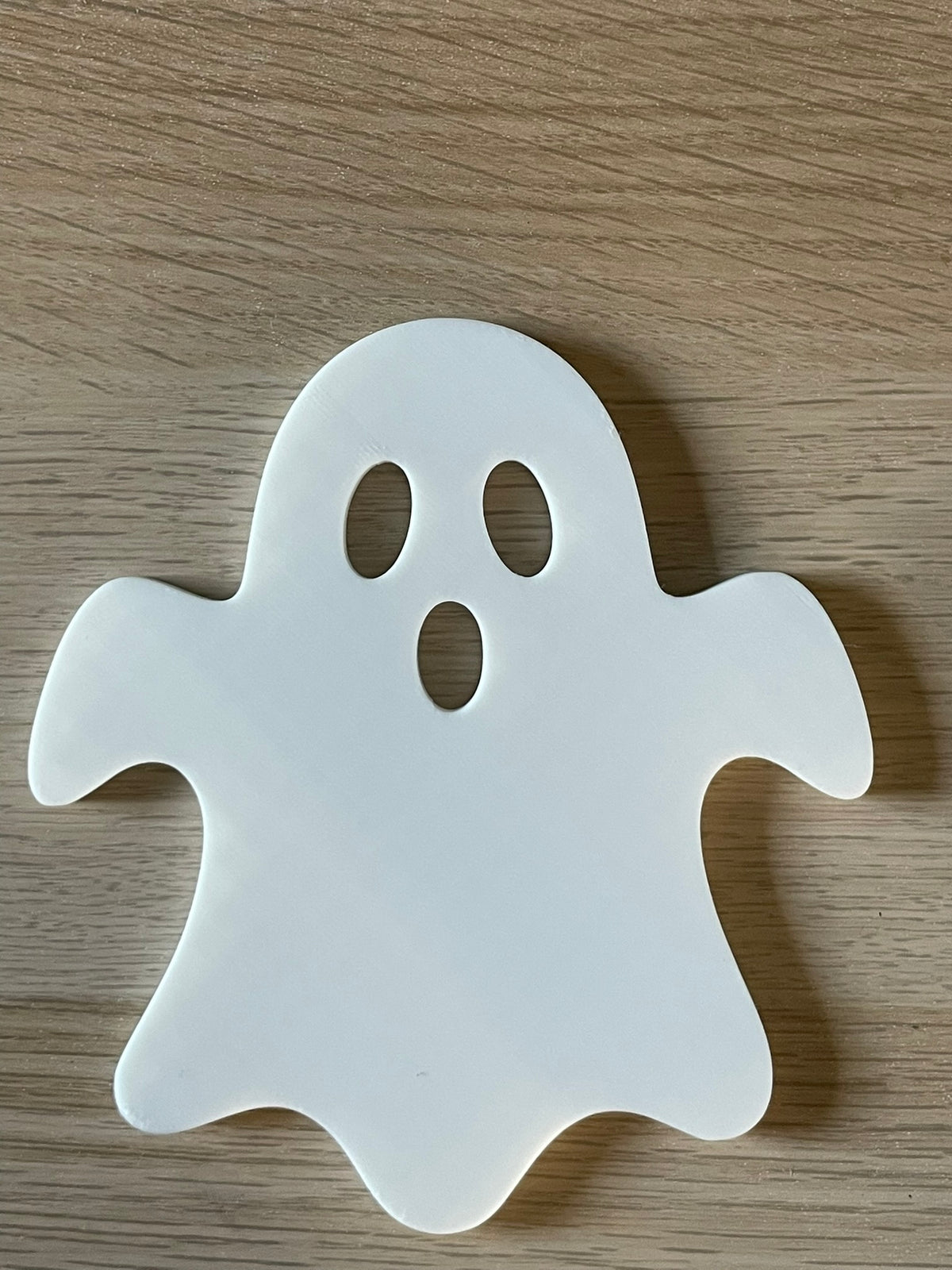 A white ghost-shaped coaster with oval and circular eye cutouts, placed on a light brown wooden surface.