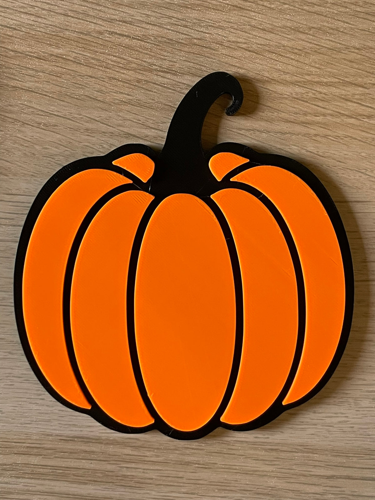 A flat orange pumpkin-shaped coaster with a black outline, placed on a light wood-grain surface.