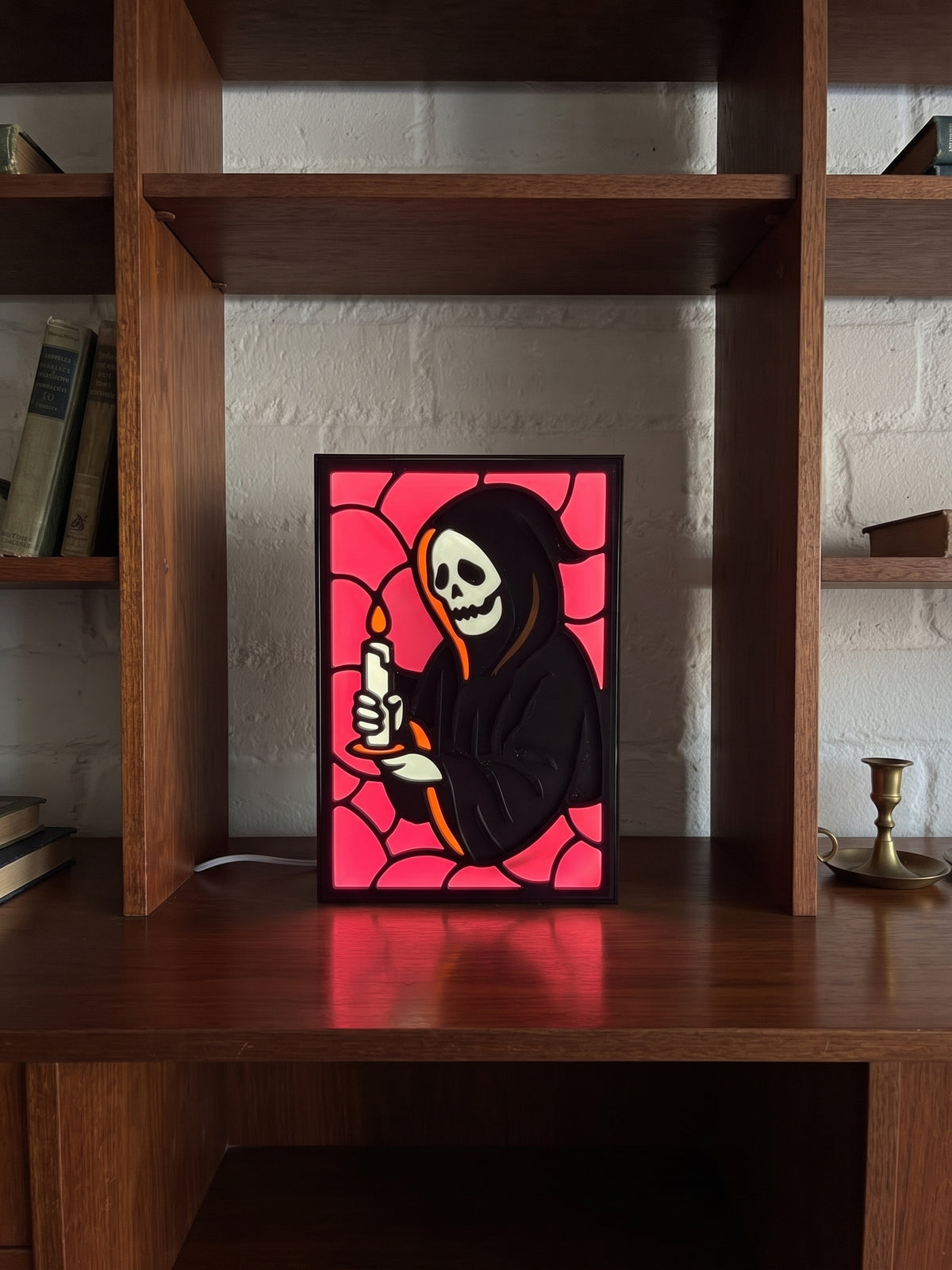 A framed stained-glass-style art print featuring a hooded skeleton holding a candle, displayed on a wooden shelf in front of a white brick wall.
