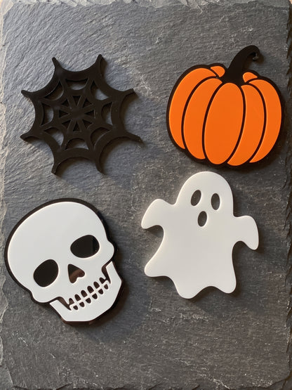 Four Halloween-themed flat decorative coasters arranged on a dark gray slate surface: a black spiderweb, an orange pumpkin, a white skull, and a white ghost.