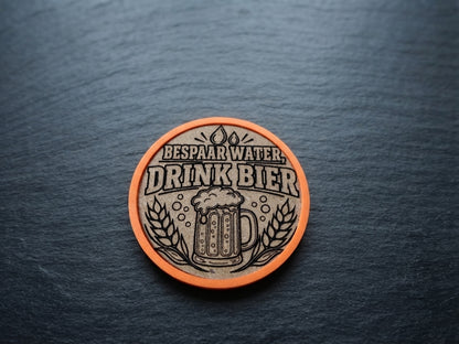 A circular cork beer token or coaster with an orange border, featuring the Dutch text 'BESPAAR WATER DRINK BIER' and an engraved beer mug with wheat, placed on a dark slate background.