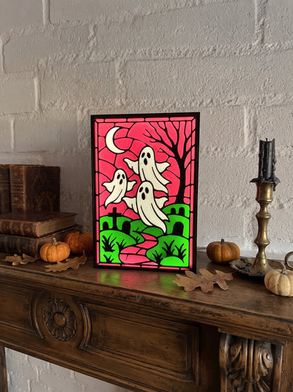 A framed Halloween art print featuring three white ghosts, a black tree, and a magenta sky with a green hill, displayed on a wooden mantel with pumpkins and books against a white brick wall.