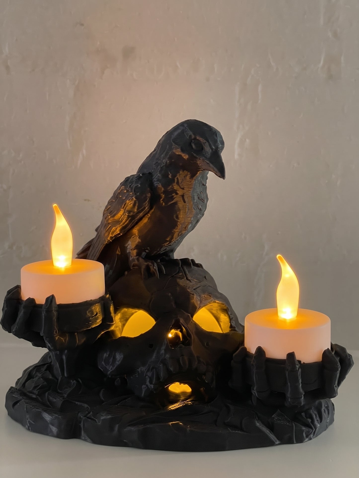 A gothic Halloween candle holder featuring a black raven perched on a skull, with two lit white pillar candles in claw-like holders on either side and one inside, set against a neutral textured wall.