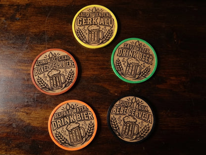 Five circular beer-themed coasters arranged on a dark wooden surface, each with a different colored rim and brown engraved label featuring a beer mug and text