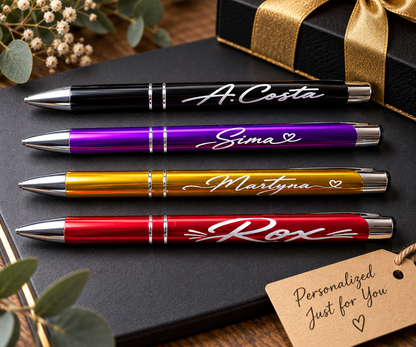 Four personalized engraved ballpoint pens in black, purple, gold, and red, each engraved with a name, heart symbol, arranged on a dark notebook next to a gift box and a 'Personalized Just for You' tag.