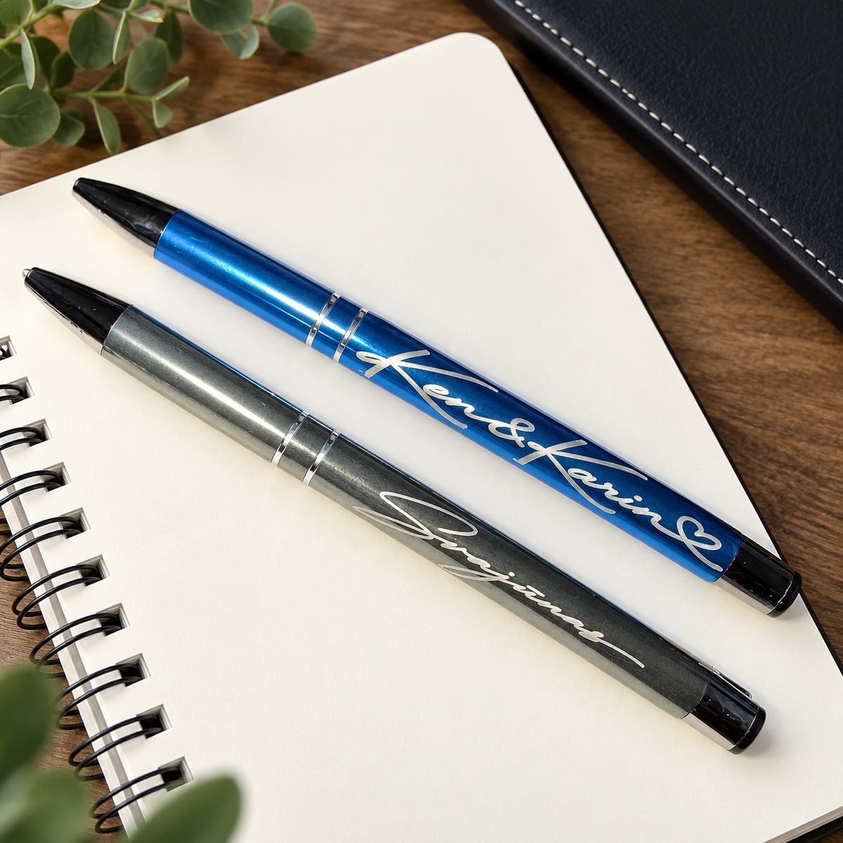 Two personalized ballpoint pens, one metallic blue and one metallic gray, engraved with the names 'Ken & Karin' and 'Svajunas', displayed on an open white spiral notebook.