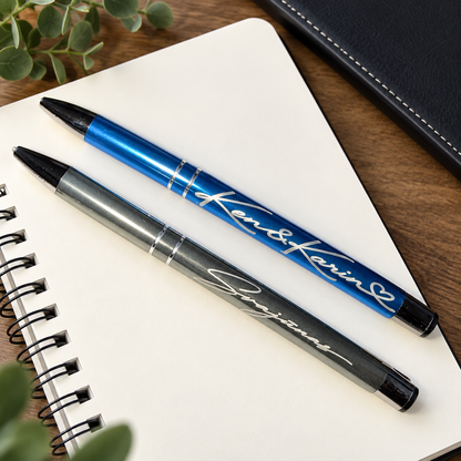 Two personalized ballpoint pens, one metallic blue and one metallic gray, engraved with the names 'Ken & Karin' and 'Svajunas', displayed on an open white spiral notebook.