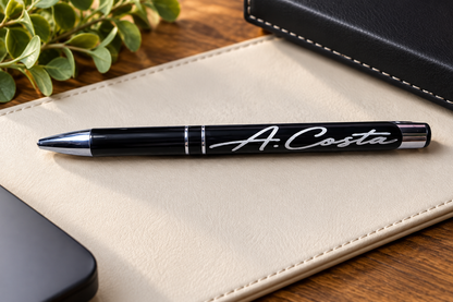A black ballpoint pen and the engraved name 'A. Costa' printed in white script, resting on a beige leather notebook cover over a wooden desk.