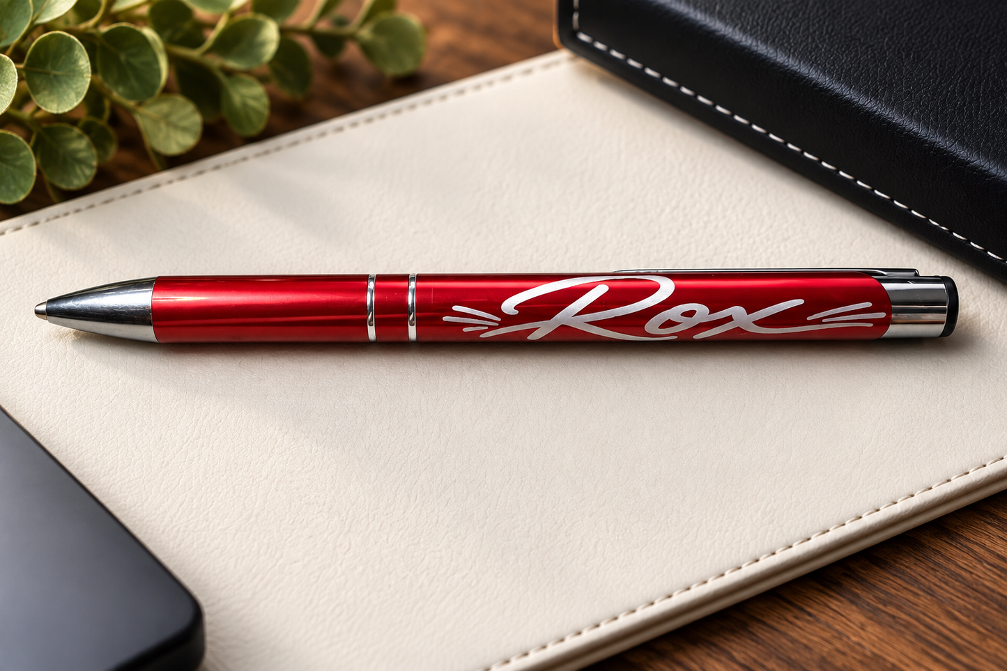 A metallic red ballpoint pen with a white engraved script reading 'Rox' on the barrel, resting on a cream-colored leather notebook cover over a wooden desk.